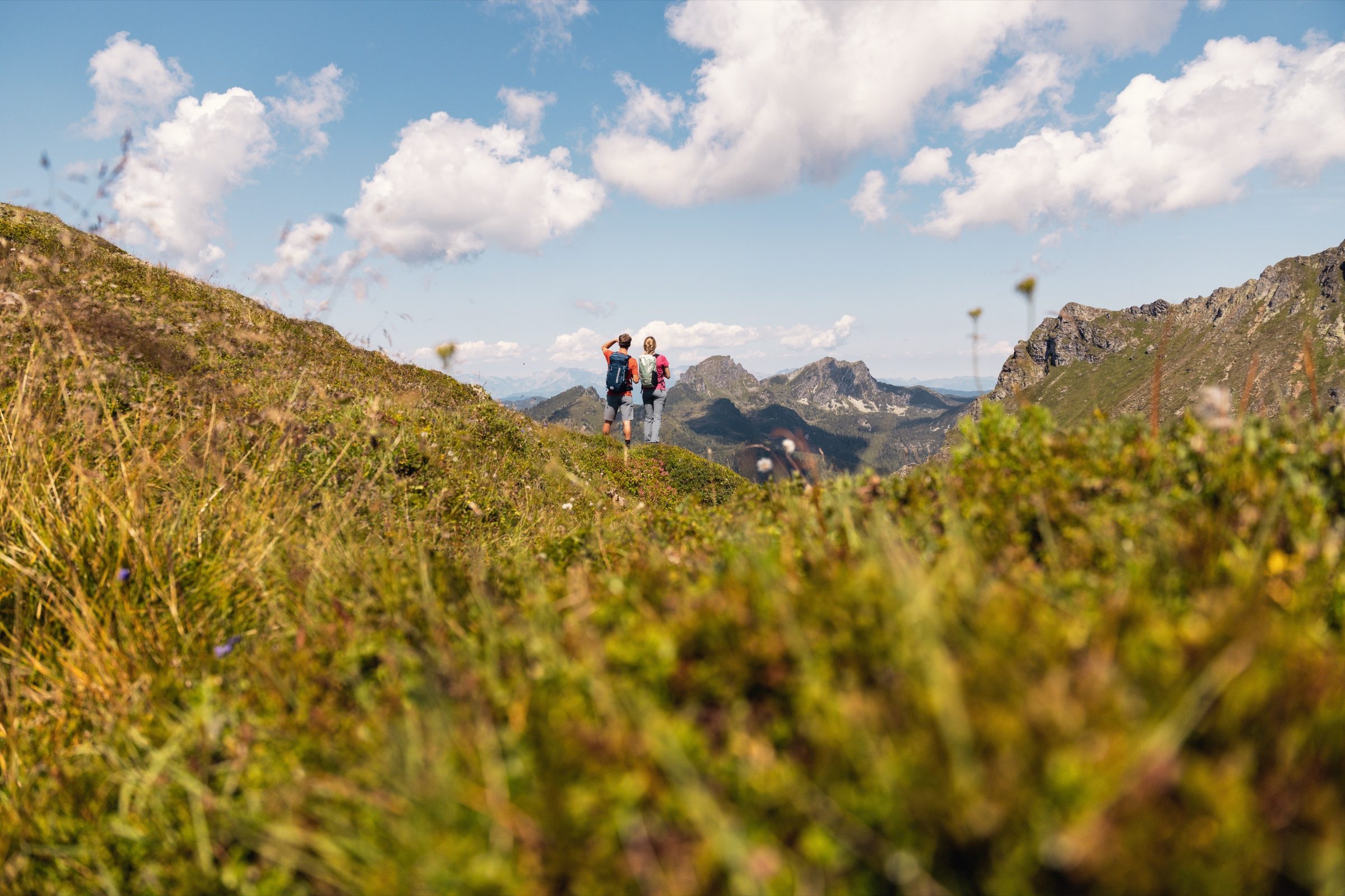 Zwei Wanderer auf einem grünen Grat vor einem weiten Bergpanorama über Kappl im Paznaun im Sommer