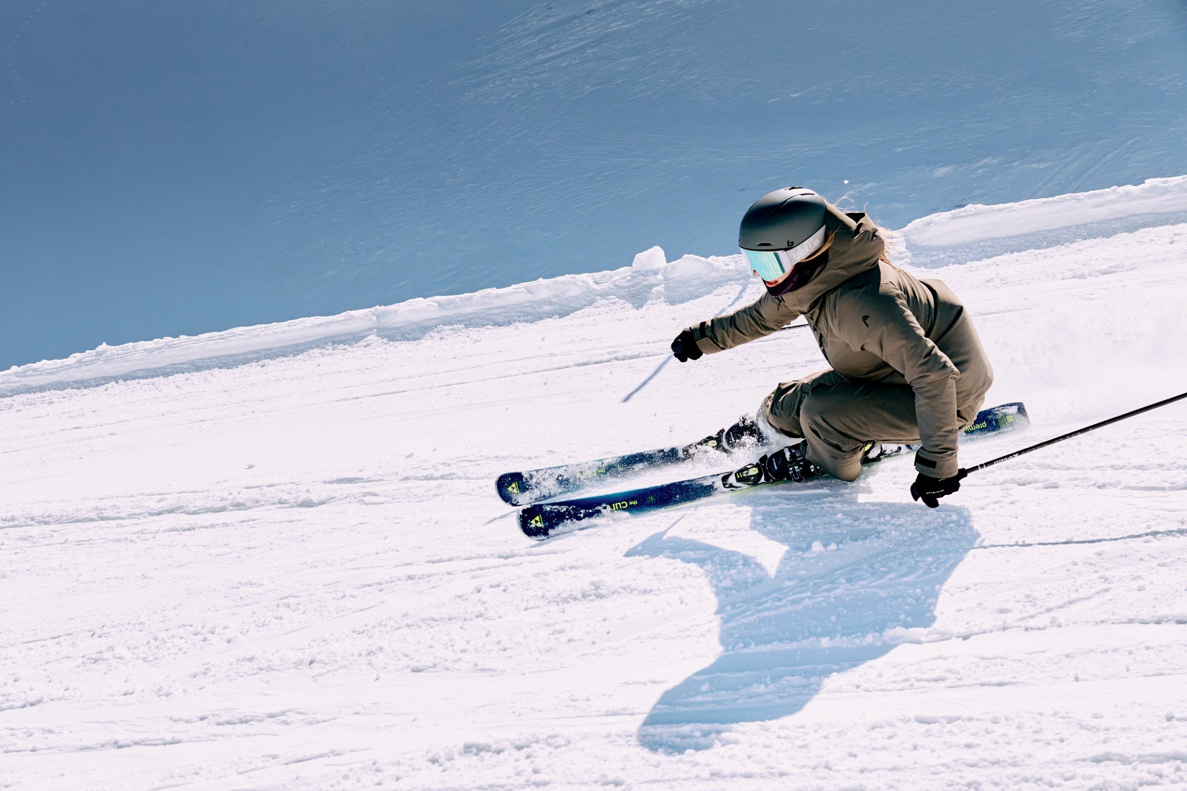 Skifahrer in tiefer Carving-Kurve auf frisch präparierter Piste, langer Schatten, tiefblauer Himmel — INTERSPORT RENT Winter 2026/27
