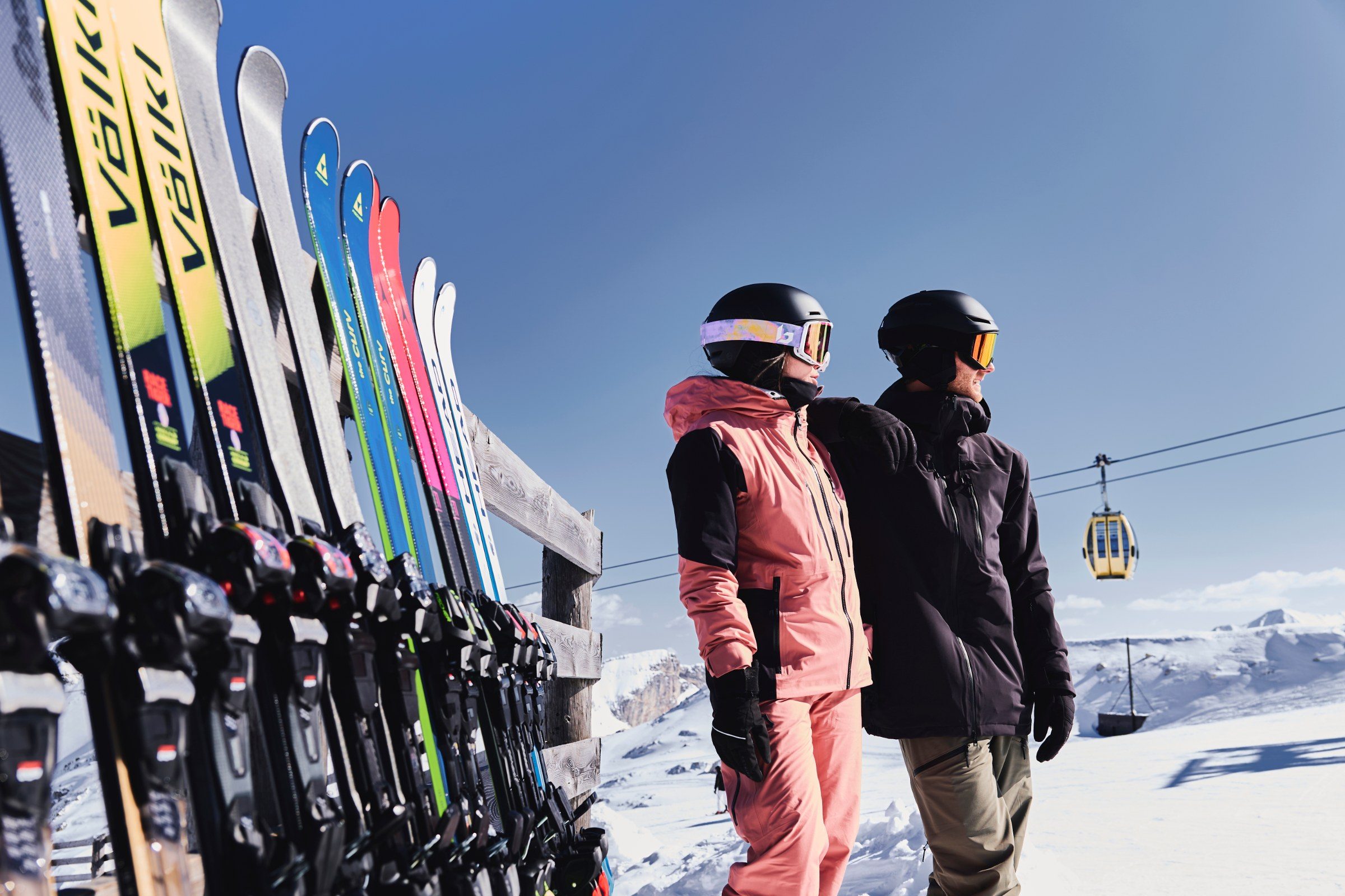 Paar in Skibekleidung vor einem Skiständer voller Verleihski, im Hintergrund die Gondelbahn — INTERSPORT RENT Winter 2026/27