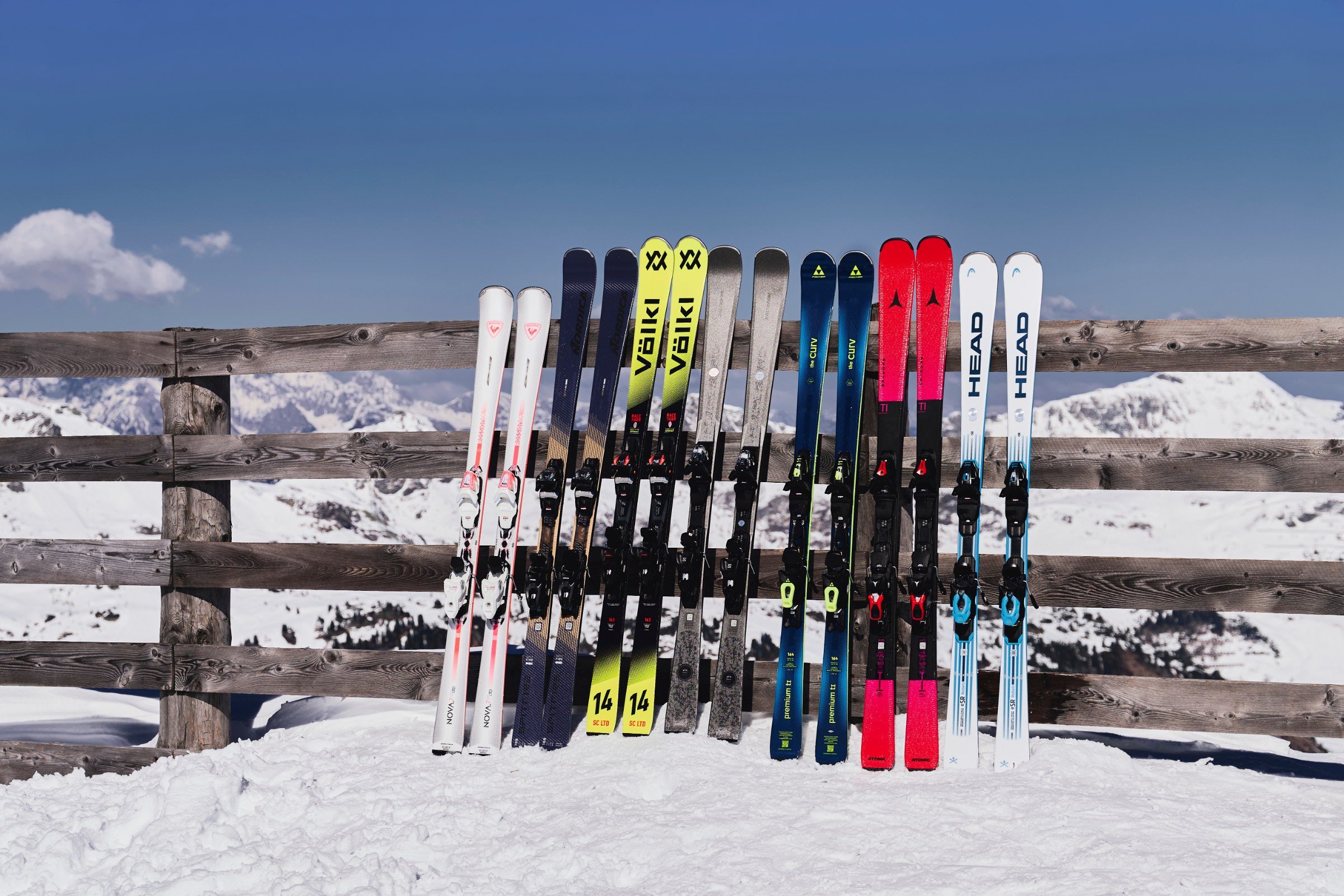 Skiständer mit Verleihski von Völkl, Head, Atomic und Rossignol an einem Holzzaun vor Bergpanorama — INTERSPORT RENT Winter 2026/27