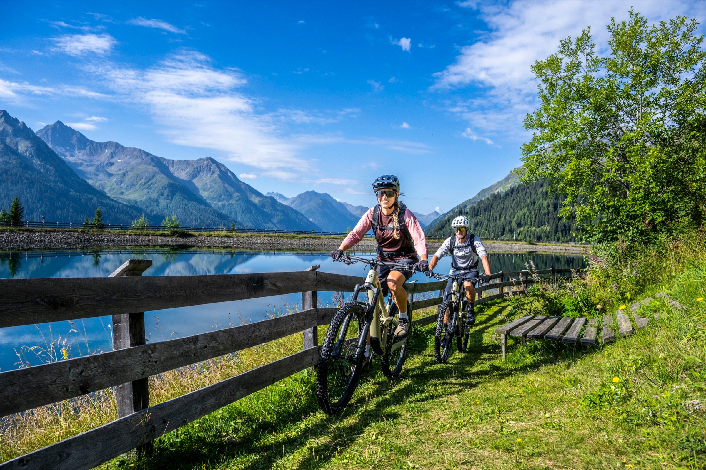 Zwei E-Mountainbiker rollen an einem Bergsee entlang, dahinter die Paznauner Gipfel