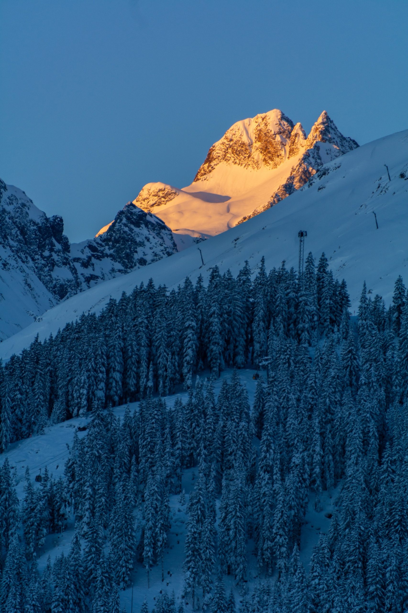 Alpenglühen auf einem Paznauner Gipfel im ersten Morgenlicht, verschneite Wälder im Schatten darunter