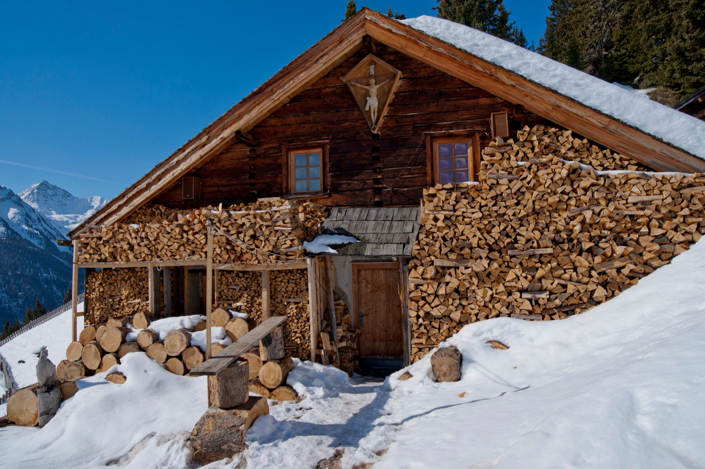 Verschneiter Tiroler Stadel mit aufgeschichteter Brennholzwand und Wegkreuz, hoch über dem Paznaun